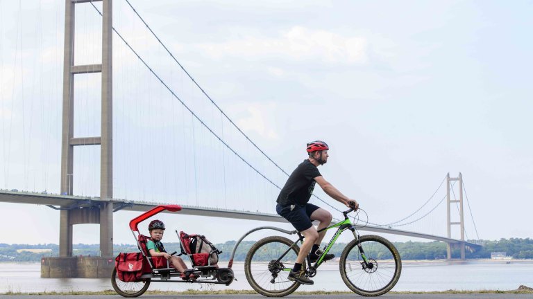 Chris Vaughan Photography - commercial photography | A dad cycles, towing his son in a cart behind him, along a cycle path with the Humber Bridge in the background.