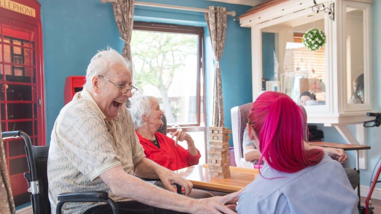 A care home resident, sat in a wheelchair, jokes with a carer.