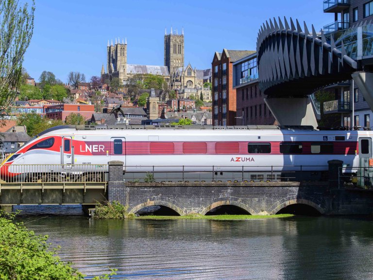 Chris Vaughan Photography - commercial photography | An LNER Azusa is pictured passing under a modern bridge in Lincoln city centre. In the foreground is water from the Brayford Pool reflecting the train, whilst Lincoln Cathedral is seen in the background.