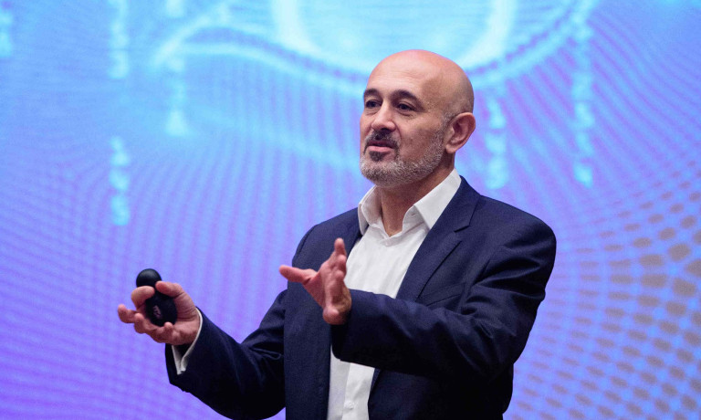 Chris Vaughan Photography - corporate event images | A speaker talks on stage in front of a digital image on the screen behind him.