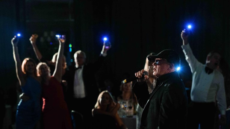 Chris Vaughan Photography - corporate event images | A performer sings as guests wave their phones, with lights on, in the background at a charity black tie dinner.