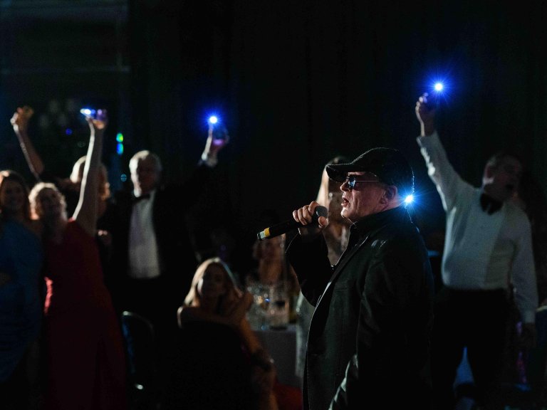 Chris Vaughan Photography - corporate event images | A performer sings as guests wave their phones, with lights on, in the background at a charity black tie dinner.