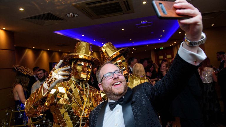 A guest takes a selfie with two entertainers, dressed in gold, at a corporate charity fundraising dinner.