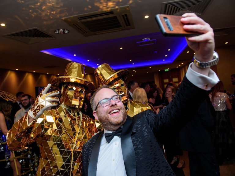 A guest takes a selfie with two entertainers, dressed in gold, at a corporate charity fundraising dinner.