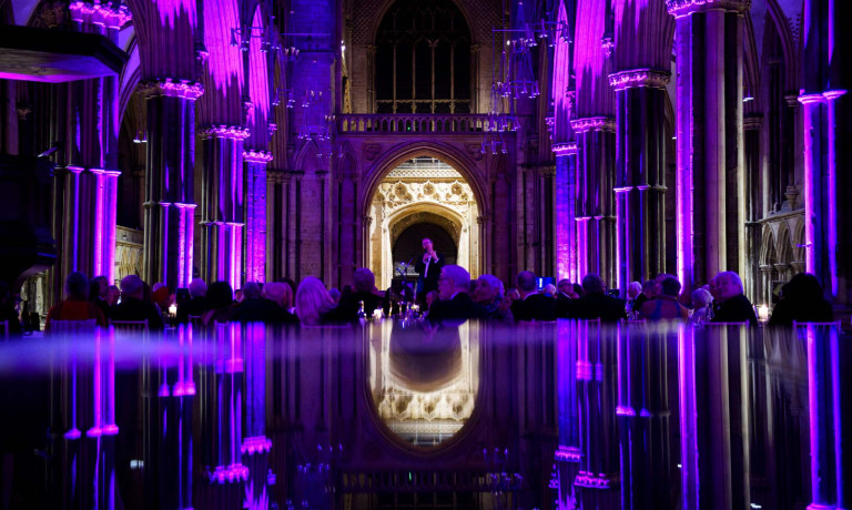 An image of Reverend Richard Coles speaking at Lincoln Cathedral. The Cathedral is illuminated purple, and the bottom half of the image is a reflection of the scene in a piano.