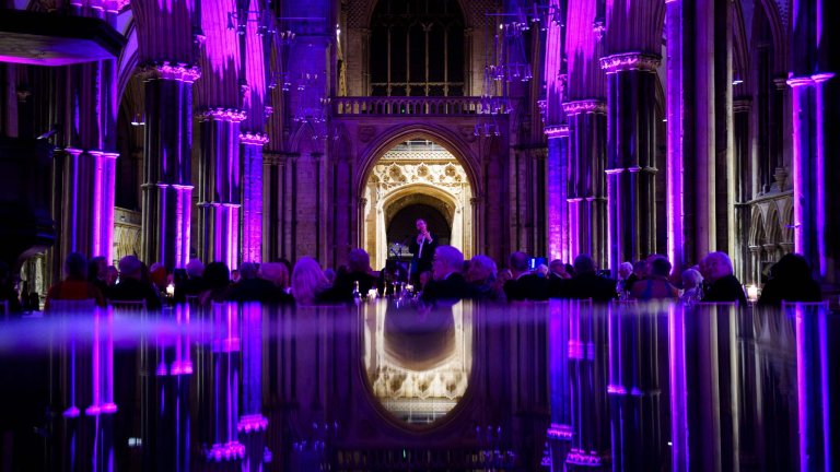 An image of Reverend Richard Coles speaking at Lincoln Cathedral. The Cathedral is illuminated purple, and the bottom half of the image is a reflection of the scene in a piano.