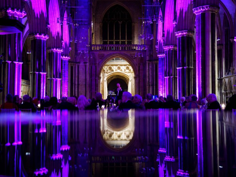 An image of Reverend Richard Coles speaking at Lincoln Cathedral. The Cathedral is illuminated purple, and the bottom half of the image is a reflection of the scene in a piano.
