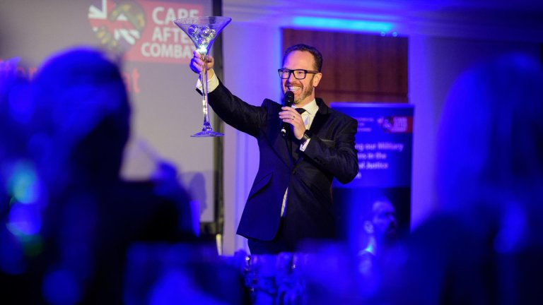 A singer performing at a corporate charity fundraising dinner holds aloft (in the style of a toast) a table centrepiece in the shape of a large glass.