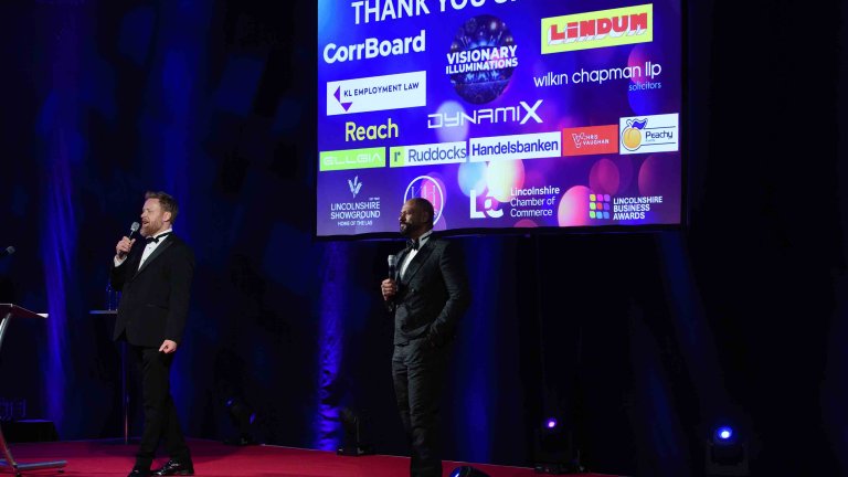 Two of the hosts on stage with some of the event sponsors logo on the screen behind them.