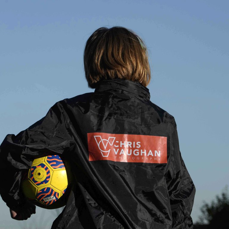 A picture of Chris' daughter wearing a new Chris Vaughan Photography sponsored rain jacket. She is looking away from the camera, showing the branding on the back. She is holding a yellow football.
