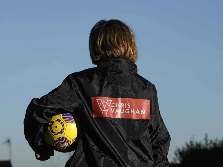 A picture of Chris' daughter wearing a new Chris Vaughan Photography sponsored rain jacket. She is looking away from the camera, showing the branding on the back. She is holding a yellow football.