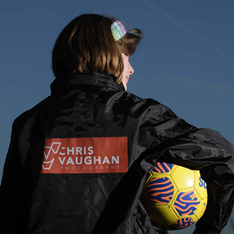 A picture of Chris' daughter wearing a new Chris Vaughan Photography sponsored rain jacket. She is looking away from the camera, showing the branding on the back, whilst looking towards her right. She is holding a yellow football.