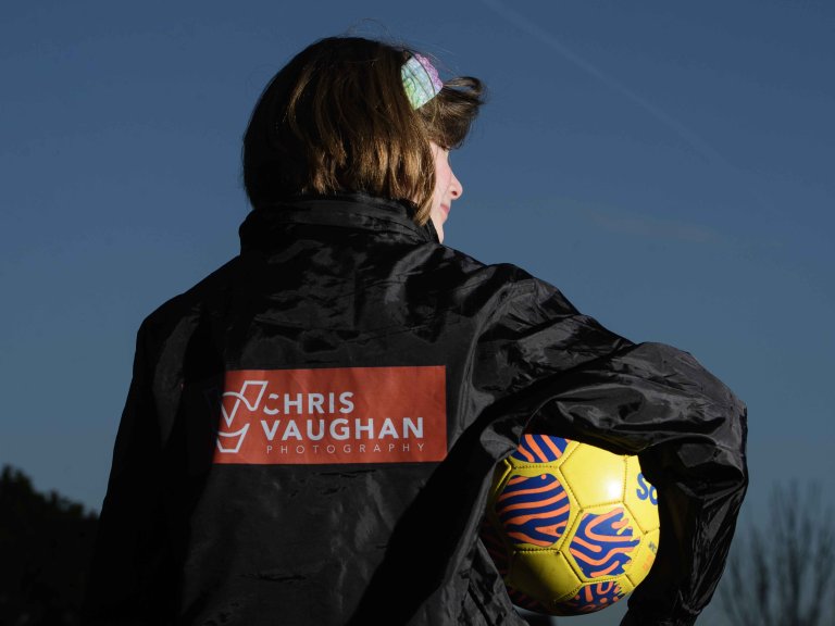 A picture of Chris' daughter wearing a new Chris Vaughan Photography sponsored rain jacket. She is looking away from the camera, showing the branding on the back, whilst looking towards her right. She is holding a yellow football.
