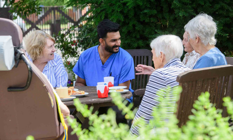 A care worker sits with a group of residents outside, as they all enjoy a cup of tea and some biscuits!.