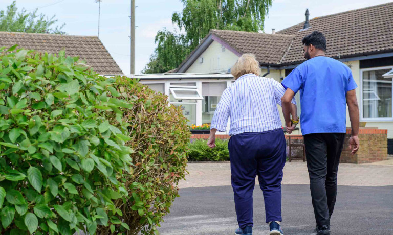 A care walker walks arm-in-arm, away from the camera, with a resident outside the care home.