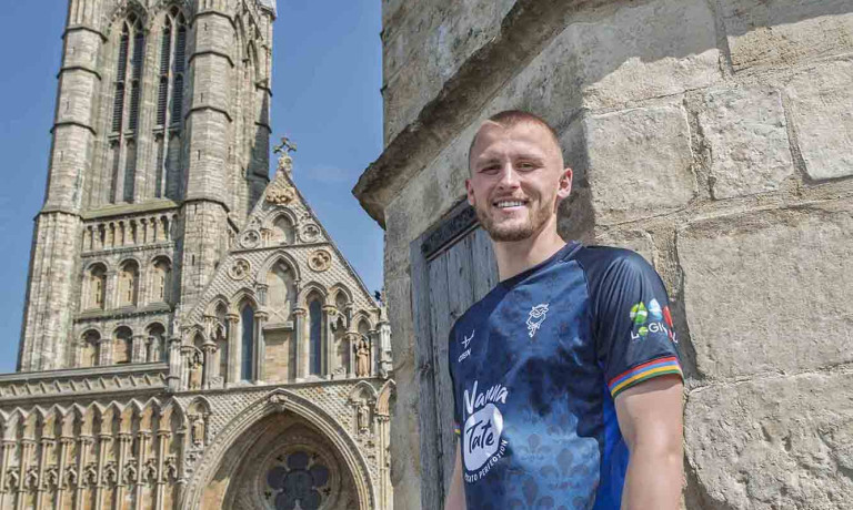 A smiling Tom Hamer of Lincoln City wearing the club's away kit for the 2025/26 season whilst stood on top of Exchequregate, with Lincoln Cathedral in the background.