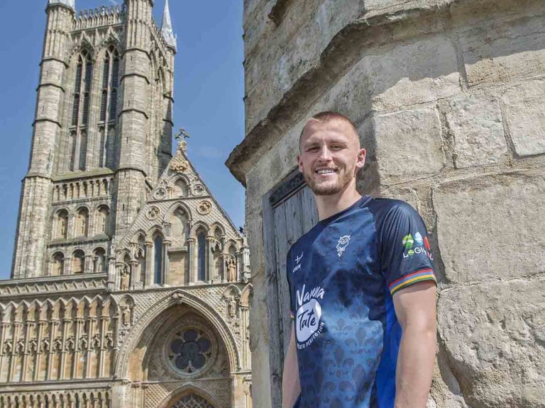 A smiling Tom Hamer of Lincoln City wearing the club's away kit for the 2025/26 season whilst stood on top of Exchequregate, with Lincoln Cathedral in the background.