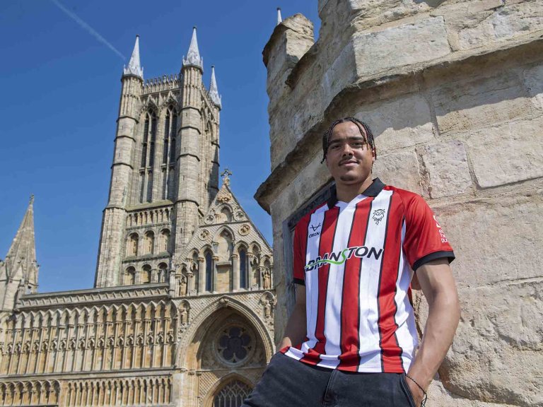 A smiling Jovon Makama of Lincoln City wearing the club's away kit for the 2025/26 season whilst stood on top of Exchequregate, with Lincoln Cathedral in the background.