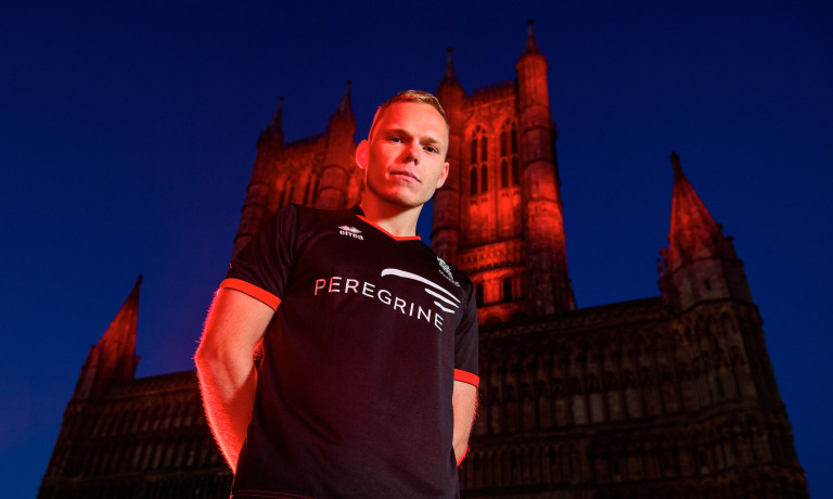 Anthony Scully wearing the 2020/21 Lincoln City black away kit with Lincoln Cathedral illuminated red behind him.