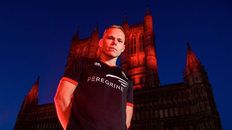 Anthony Scully wearing the 2020/21 Lincoln City black away kit with Lincoln Cathedral illuminated red behind him.