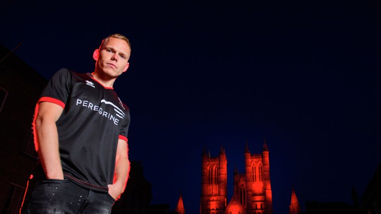 Anthony Scully wearing the 2020/21 Lincoln City black away kit with Lincoln Cathedral illuminated red behind him.