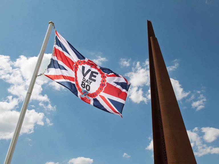 A Union flag with the VE Day 80 logo in the middle flying with the spire monument at International Bomber Command in the background.
