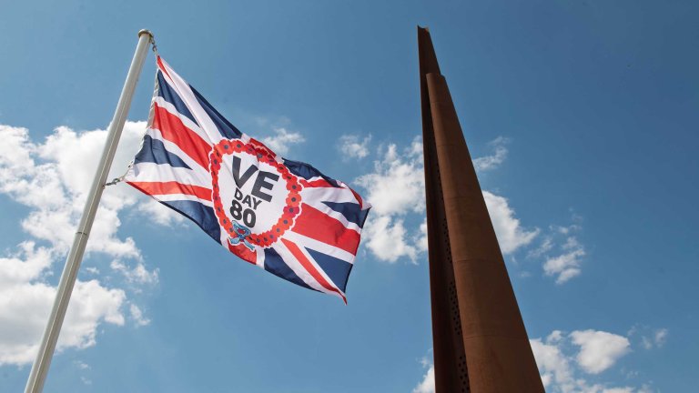 A Union flag with the VE Day 80 logo in the middle flying with the spire monument at International Bomber Command in the background.