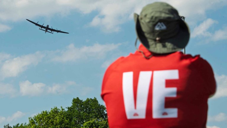 A person wearing a rugby top with VE on the back take a photograph of a Lancaster Bomber flypast.