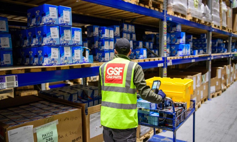 A member of the GSF Car Parts warehouse team walks amongst the aisles picking items. He is walking away from the camera, wearing a yellow high-vis jacket that shows the company name.