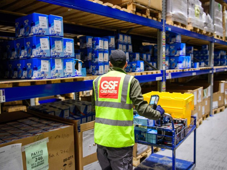 A member of the GSF Car Parts warehouse team walks amongst the aisles picking items. He is walking away from the camera, wearing a yellow high-vis jacket that shows the company name.