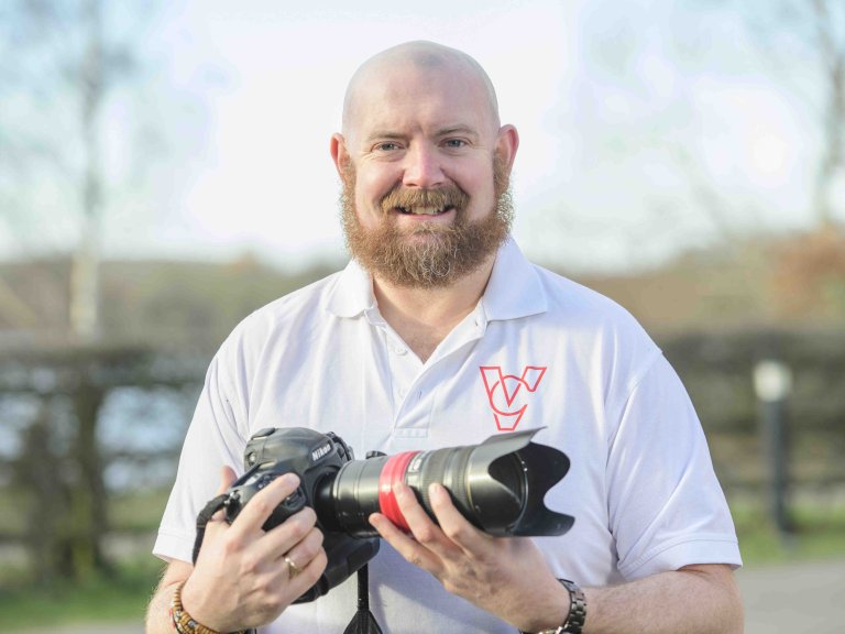 A profile picture of Chris Vaughan, owner of Chris Vaughan Photography, holding a camera and looking directly at the camera. Chris is wearing a white branded t-shirt.