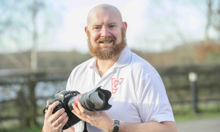 A profile picture of Chris Vaughan, owner of Chris Vaughan Photography, holding a camera and looking directly at the camera. Chris is wearing a white branded t-shirt.