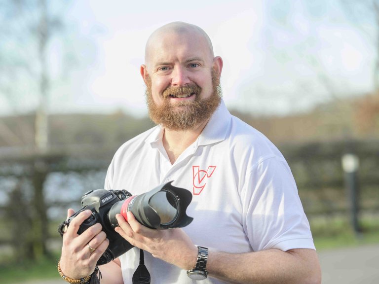A profile picture of Chris Vaughan, owner of Chris Vaughan Photography, holding a camera and looking directly at the camera. Chris is wearing a white branded t-shirt.