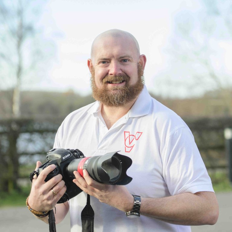 A profile picture of Chris Vaughan, owner of Chris Vaughan Photography, holding a camera and looking directly at the camera. Chris is wearing a white branded t-shirt.