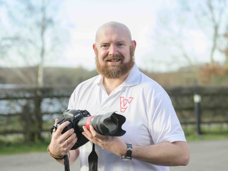 A profile picture of Chris Vaughan, owner of Chris Vaughan Photography, holding a camera and looking directly at the camera. Chris is wearing a white branded t-shirt.