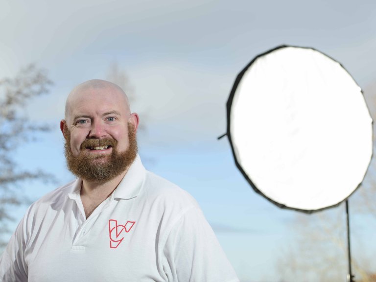 A profile picture of Chris Vaughan, owner of Chris Vaughan Photography looking directly at the camera. Chris is wearing a white branded t-shirt. Behind his is a portable studio light.