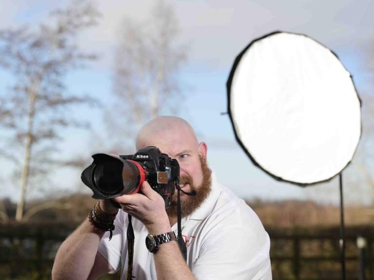 A profile picture of Chris Vaughan, owner of Chris Vaughan Photography taking a photograph. Chris is wearing a white branded t-shirt. Behind his is a portable studio light.