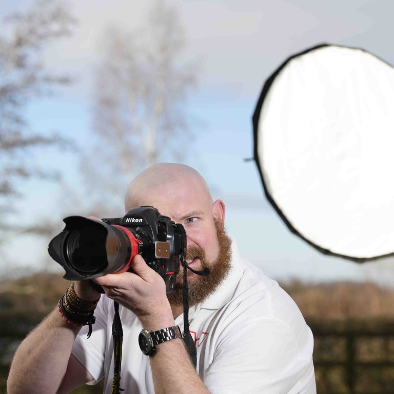 A profile picture of Chris Vaughan, owner of Chris Vaughan Photography taking a photograph. Chris is wearing a white branded t-shirt. Behind his is a portable studio light.