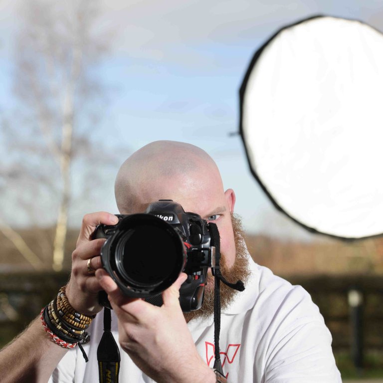 A profile picture of Chris Vaughan, owner of Chris Vaughan Photography taking a photograph. Chris is wearing a white branded t-shirt. Behind him is a portable studio light.