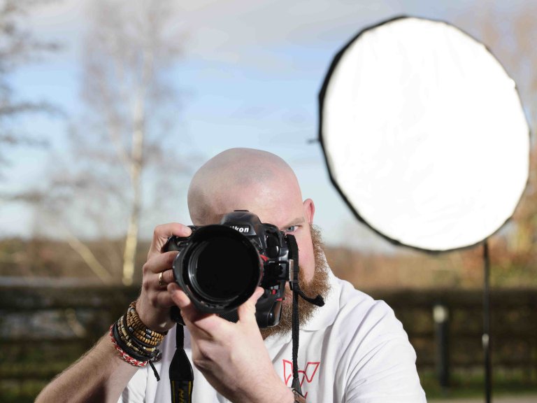 A profile picture of Chris Vaughan, owner of Chris Vaughan Photography taking a photograph. Chris is wearing a white branded t-shirt. Behind him is a portable studio light.