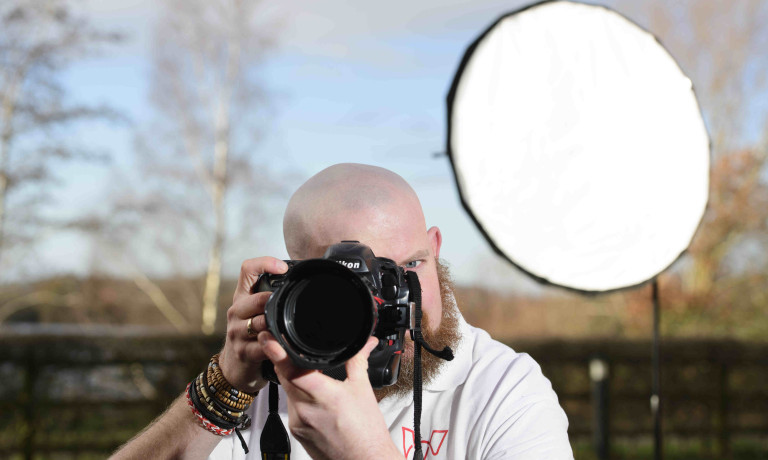A profile picture of Chris Vaughan, owner of Chris Vaughan Photography taking a photograph. Chris is wearing a white branded t-shirt. Behind him is a portable studio light.