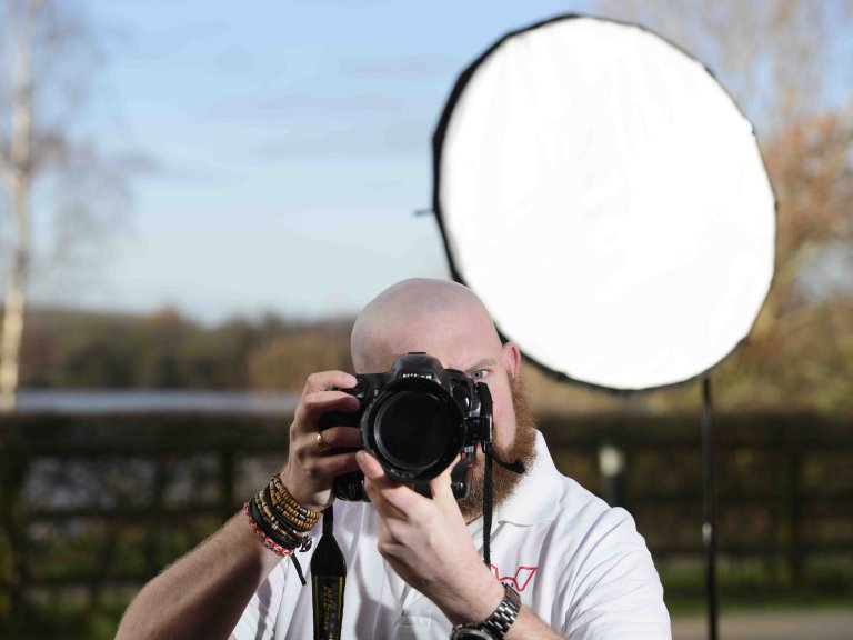 A profile picture of Chris Vaughan, owner of Chris Vaughan Photography taking a photograph. Chris is wearing a white branded t-shirt. Behind his is a portable studio light.