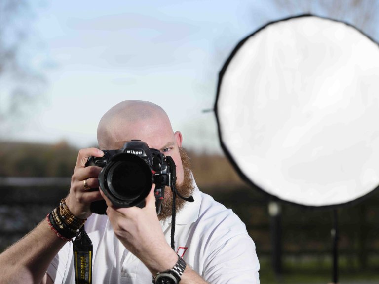 A profile picture of Chris Vaughan, owner of Chris Vaughan Photography taking a photograph. Chris is wearing a white branded t-shirt. Behind his is a portable studio light.