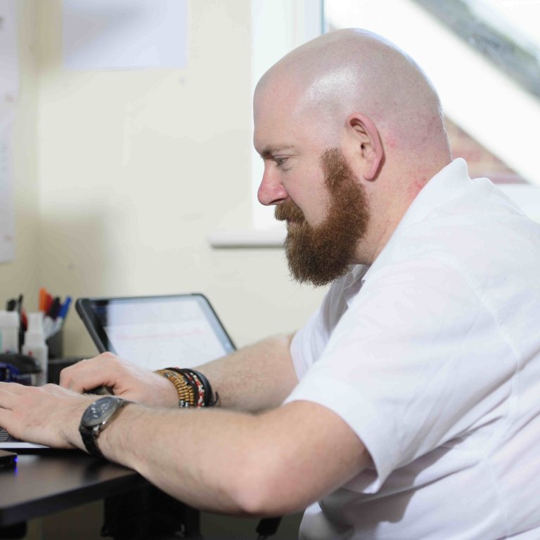 A profile picture of Chris Vaughan, owner of Chris Vaughan Photography working at his desk. Chris is wearing a white branded t-shirt.