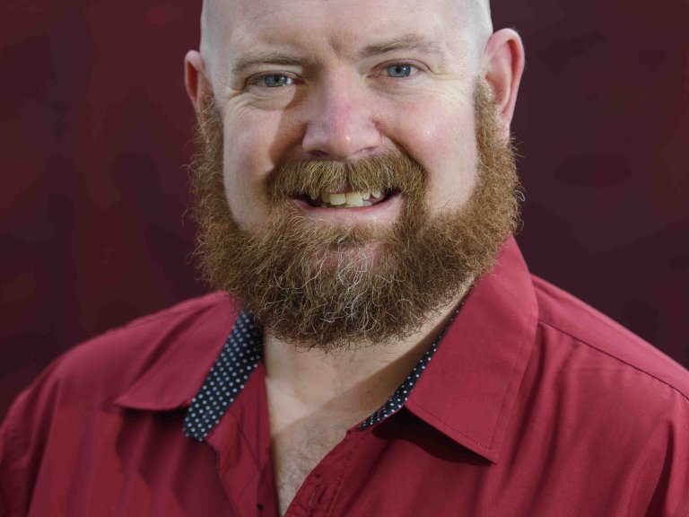 A profile picture of Chris Vaughan, owner of Chris Vaughan Photography. Chris is wearing a red shirt, and is photographed near a red brick wall.