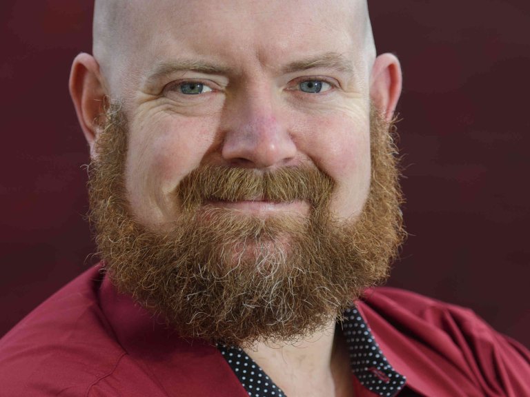 A profile picture of Chris Vaughan, owner of Chris Vaughan Photography. Chris is wearing a red shirt, and is photographed near a red brick wall.
