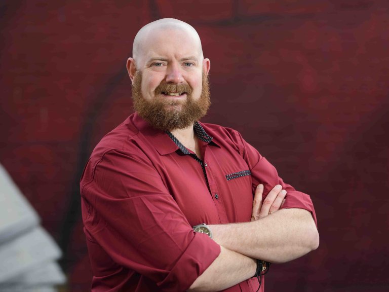 A profile picture of Chris Vaughan, owner of Chris Vaughan Photography. Chris is wearing a red shirt, and is photographed near a red brick wall.
