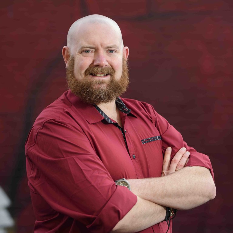 A profile picture of Chris Vaughan, owner of Chris Vaughan Photography. Chris is wearing a red shirt, and is photographed near a red brick wall.