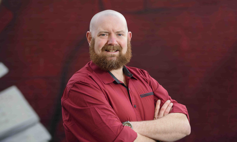 A profile picture of Chris Vaughan, owner of Chris Vaughan Photography. Chris is wearing a red shirt, and is photographed near a red brick wall.