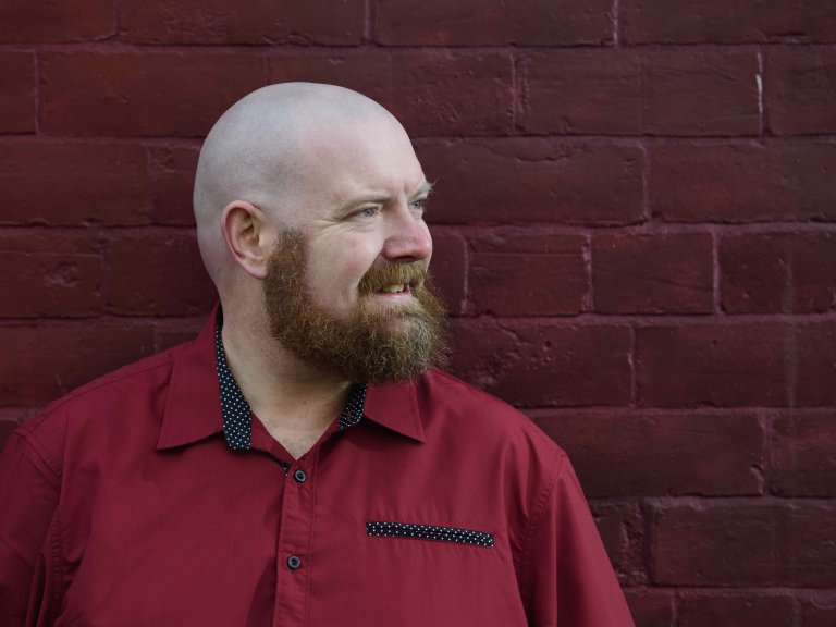 A profile picture of Chris Vaughan, owner of Chris Vaughan Photography. Chris is wearing a red shirt, and is photographed near a red brick wall.